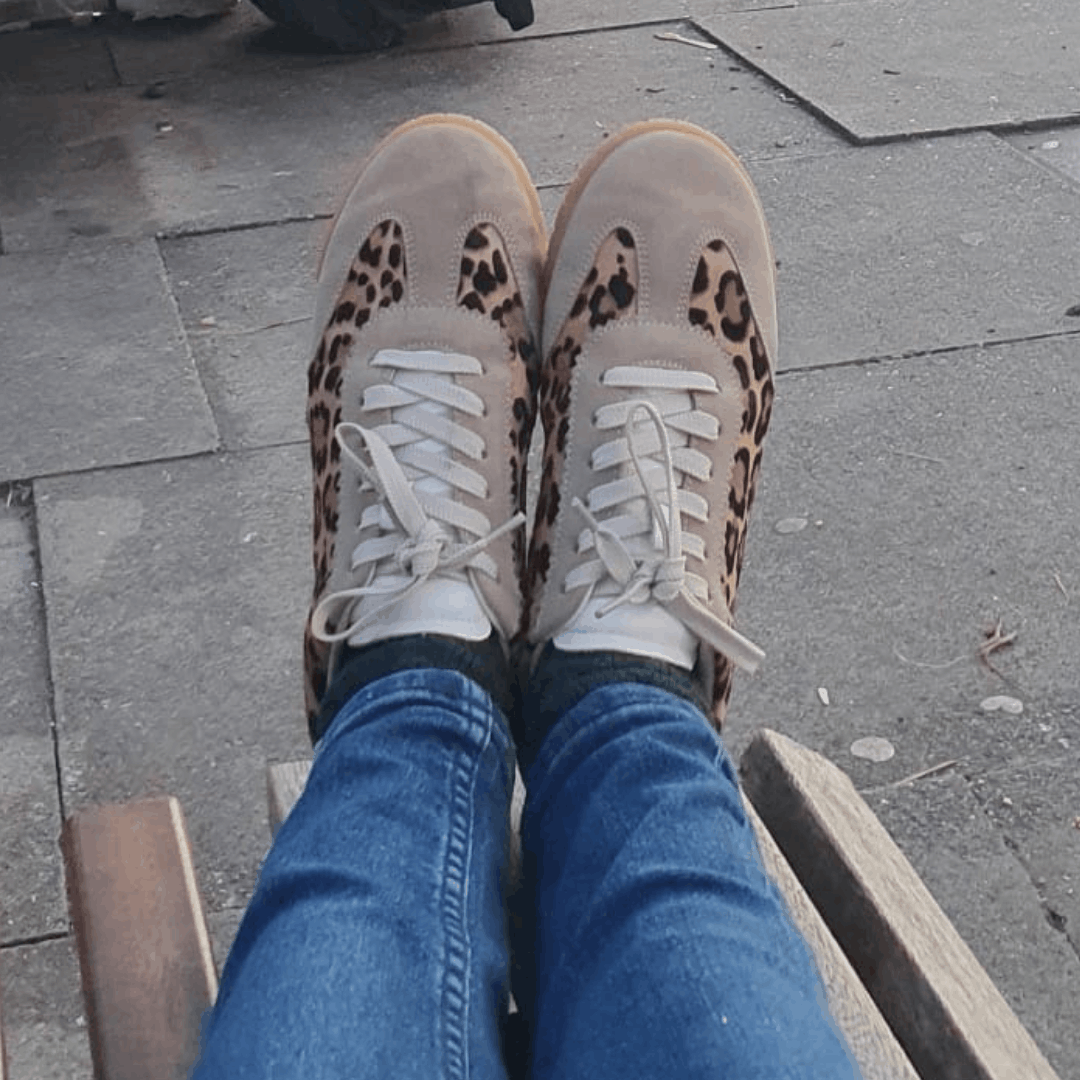 Point-of-view shot of beige suede trainers with leopard print panels and white laces worn with blue jeans on a paved walkway.