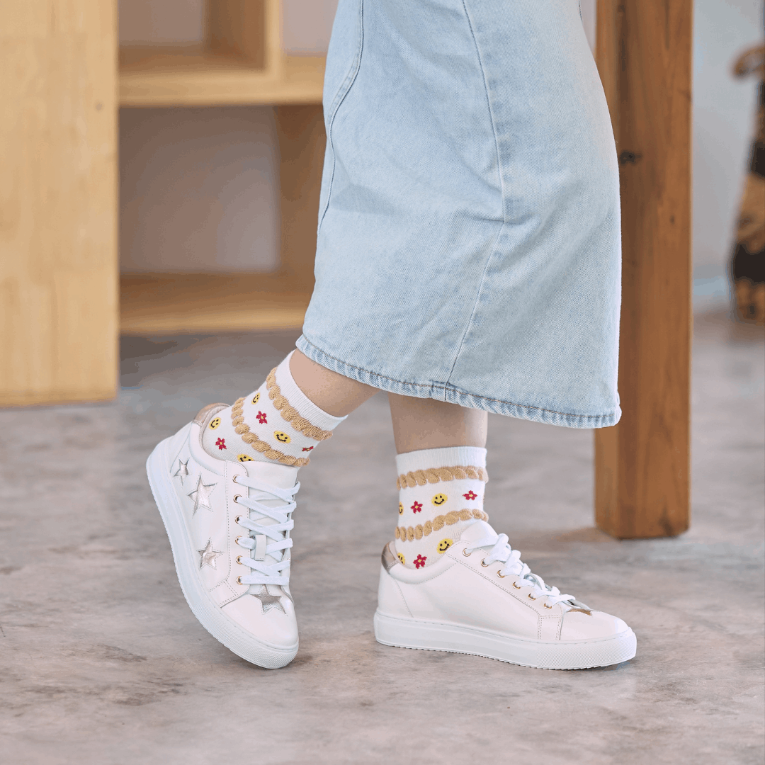 Side view of a person standing wearing white leather trainers with metallic gold star details on the outer side, paired with pattern socks and a denim skirt.