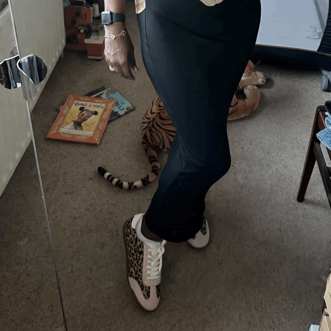 A person wearing black leggings and pink leopard print leather trainers while standing in a room with a toy tiger on the floor.