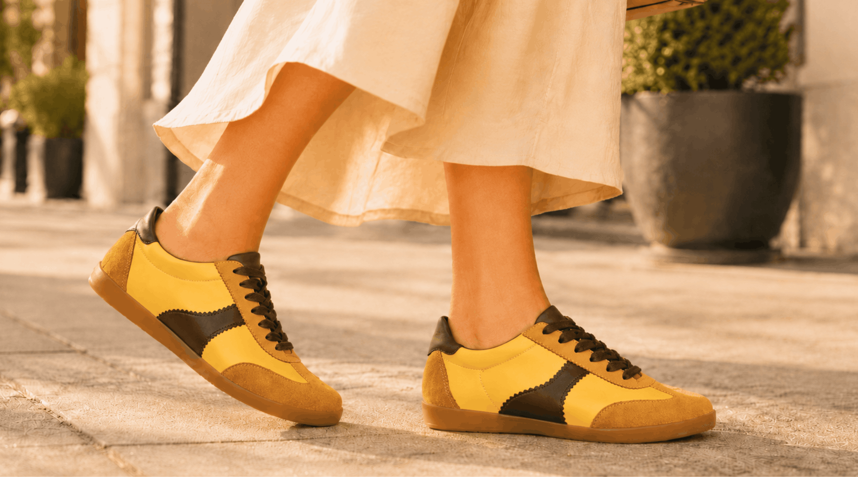 Side view of a person walking down a sunlit city street wearing Moorgate yellow and chocolate brown sneakers with a flowing cream skirt.