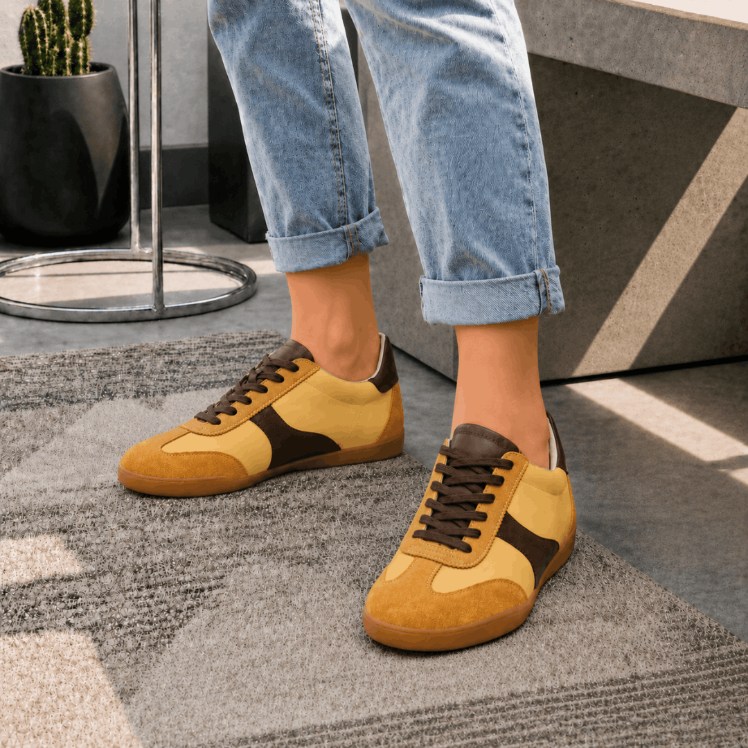 A person standing on a textured gray rug wearing cuffed blue jeans and a pair of yellow and chocolate brown leather trainers, shown from a front-quarter angle.