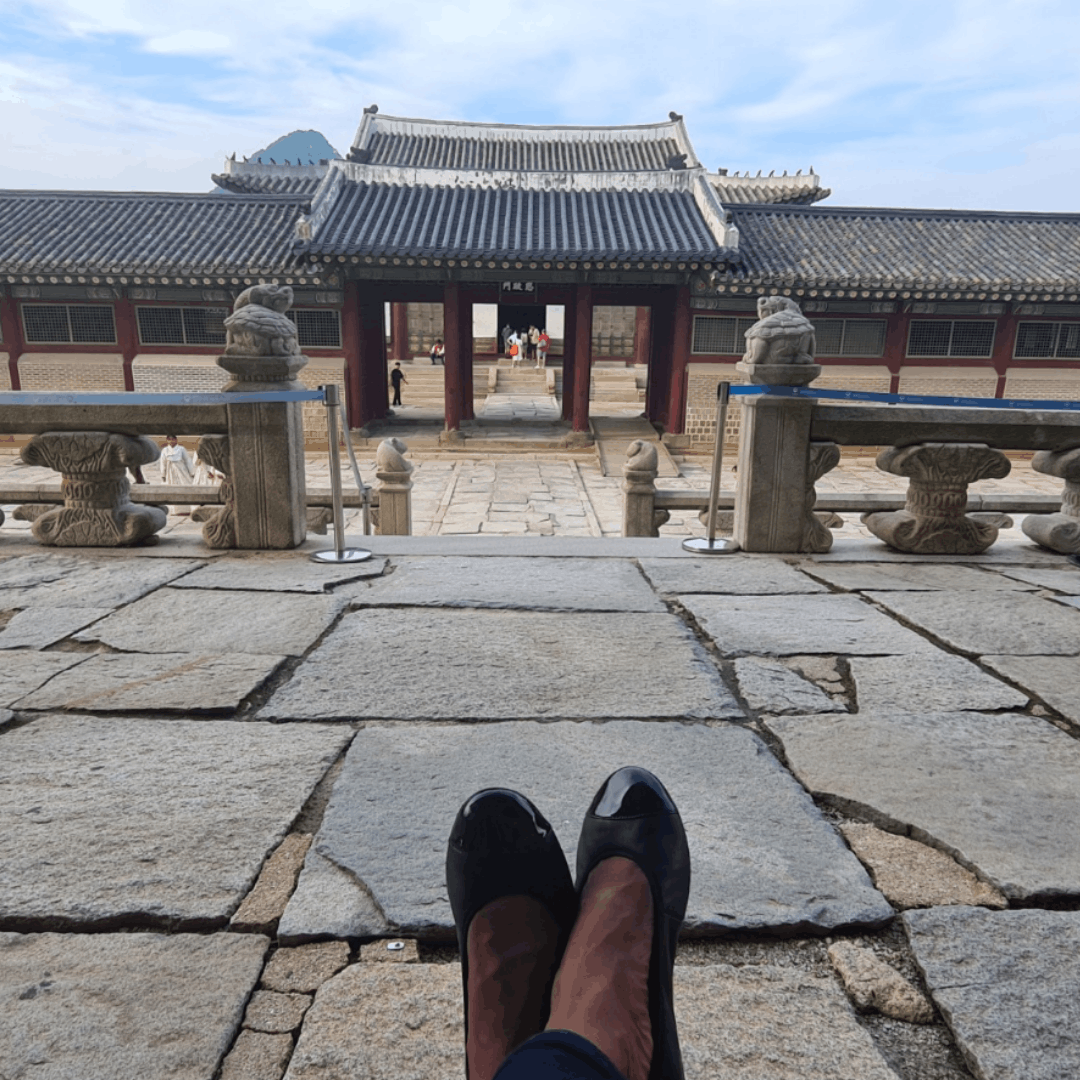High-angle, top-down view of black leather pointed-toe ballet flats resting on historic stone paving blocks in front of a traditional temple gate.