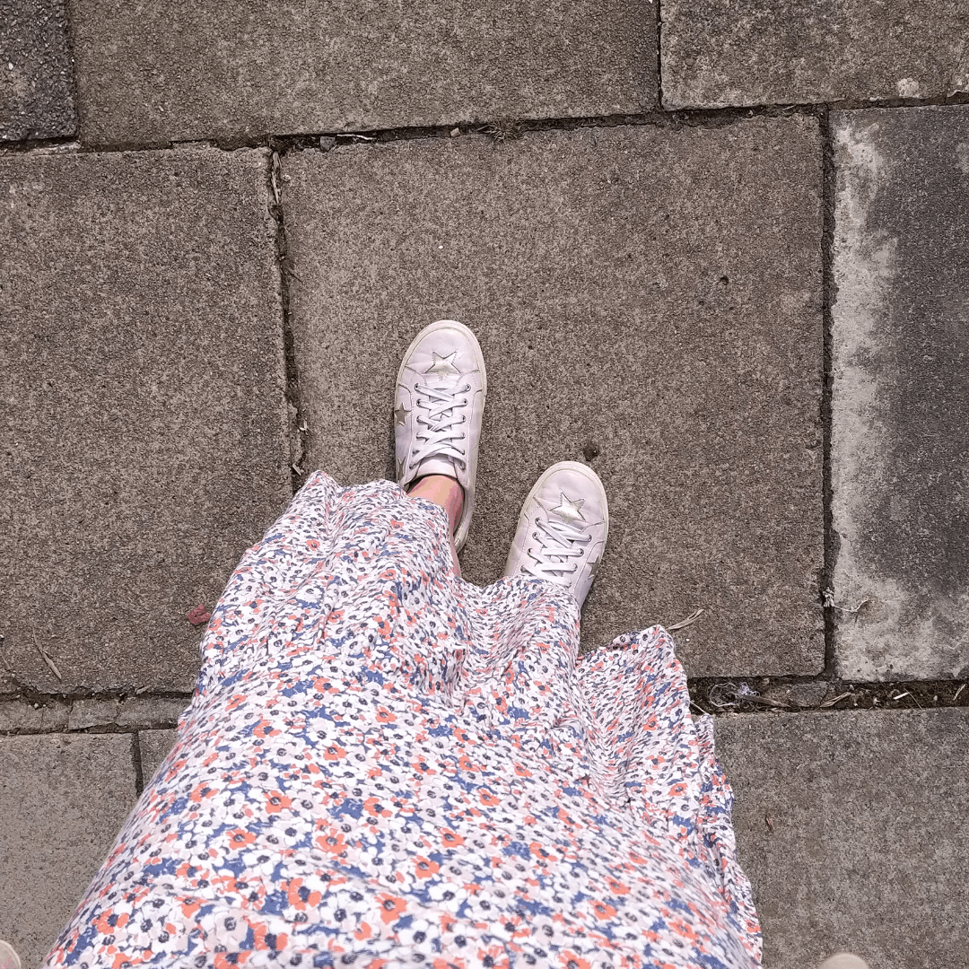 Top-down perspective of white leather lace-up sneakers with gold star accents standing on grey concrete pavement, paired with a floral print dress.