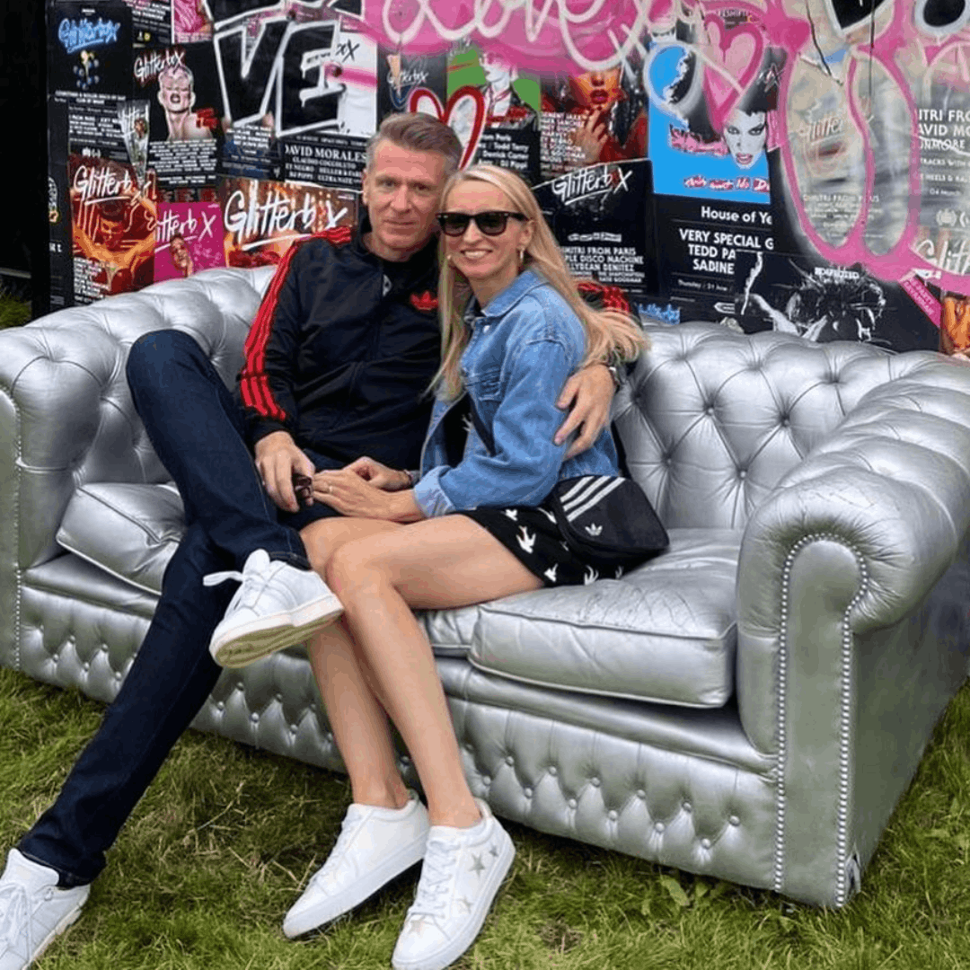 A smiling couple poses together on a silver tufted Chesterfield sofa at an outdoor event, with the woman wearing white Hoxton trainers featuring gold stars.
