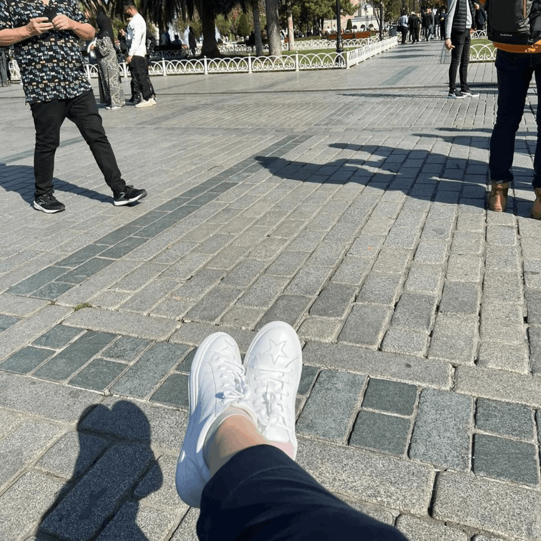 A first-person perspective shows a person relaxing outdoors on a stone-paved walkway while wearing clean, white leather Hoxton trainers with stitched star details.