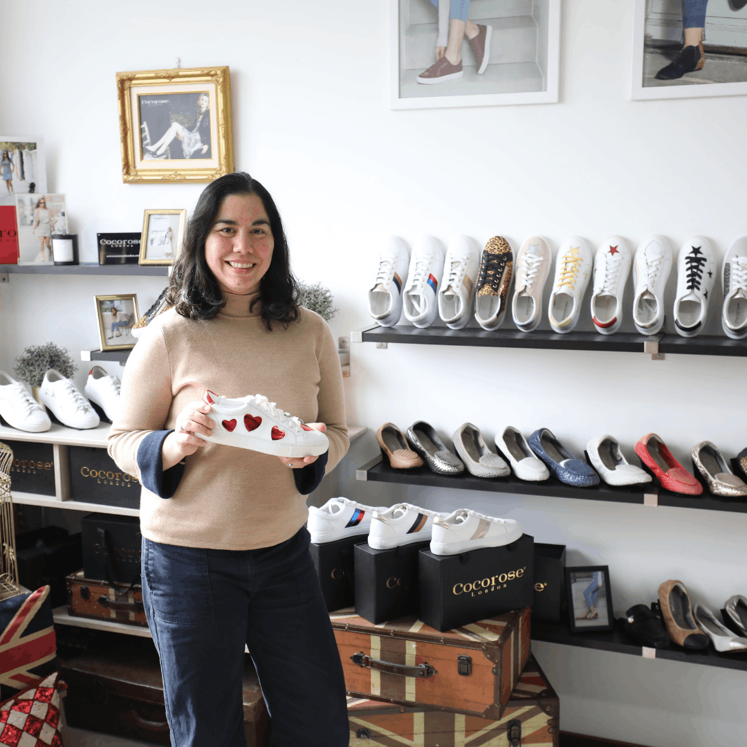 Founder Janan holding a white leather sneaker with red heart details in front of shelves filled with Cocorose London footwear.