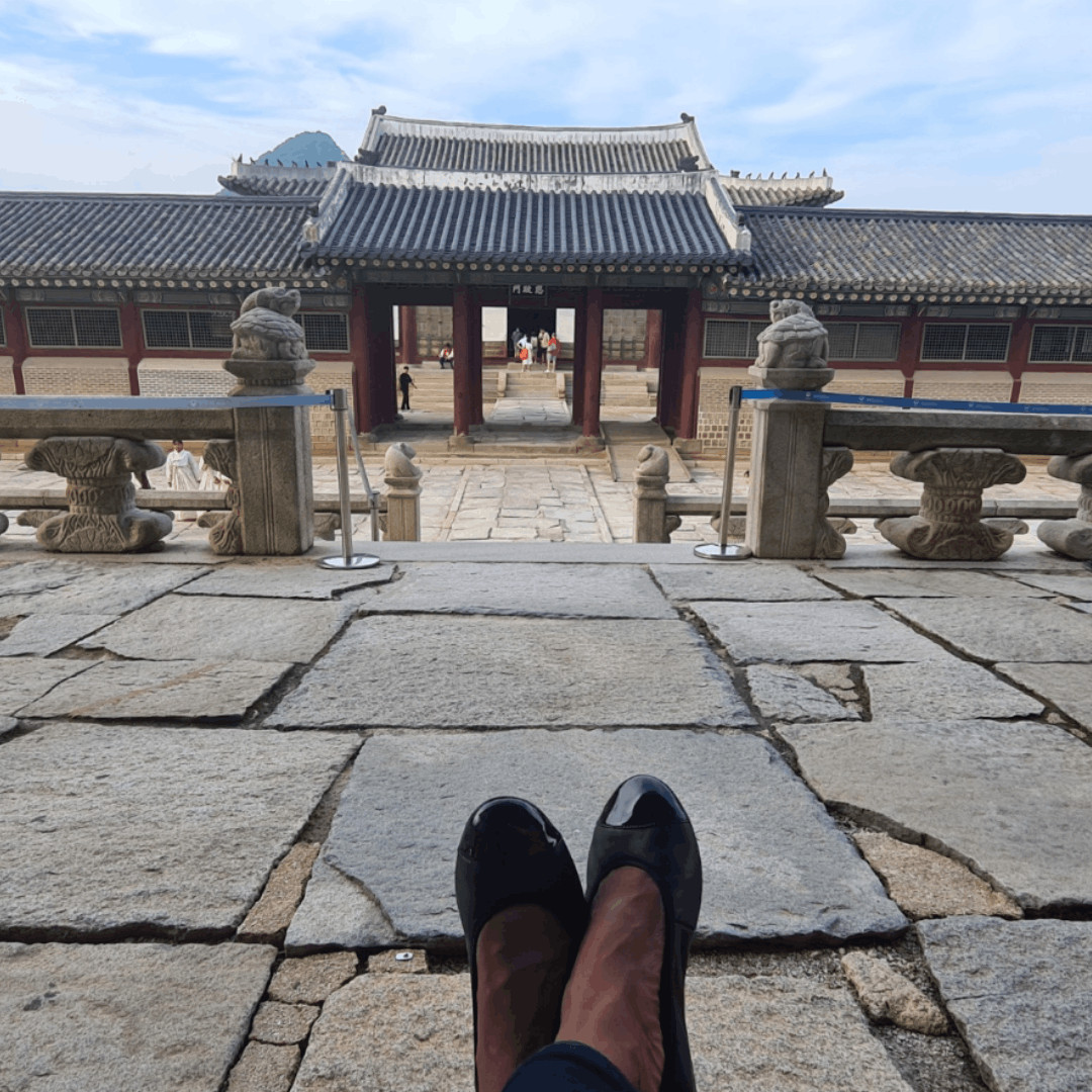 Point-of-view shot of black leather ballet flats with patent toe caps, looking toward a traditional Asian palace courtyard.
