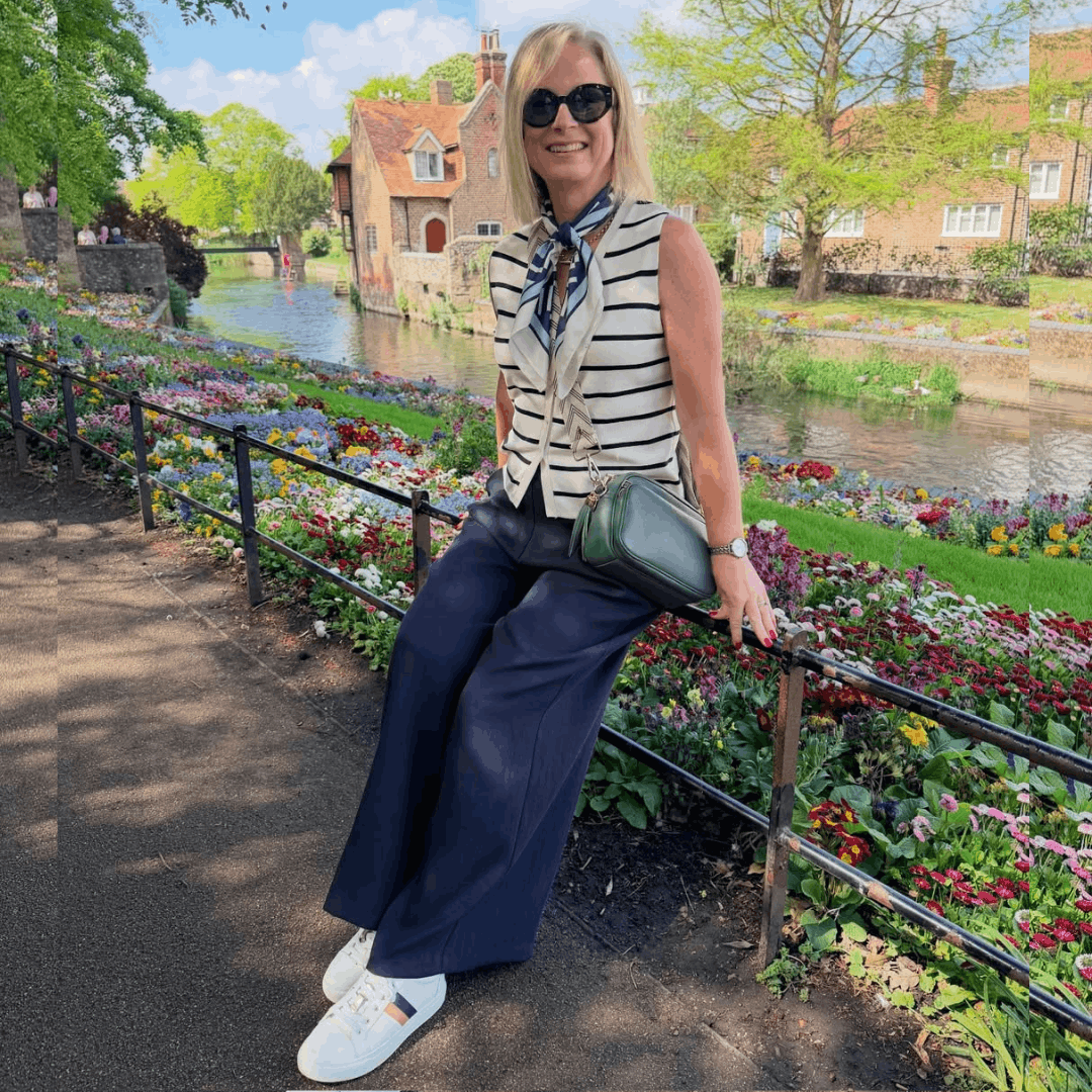 A woman sitting on a park railing by a canal wearing wide-leg navy trousers and white leather trainers with stripe details.