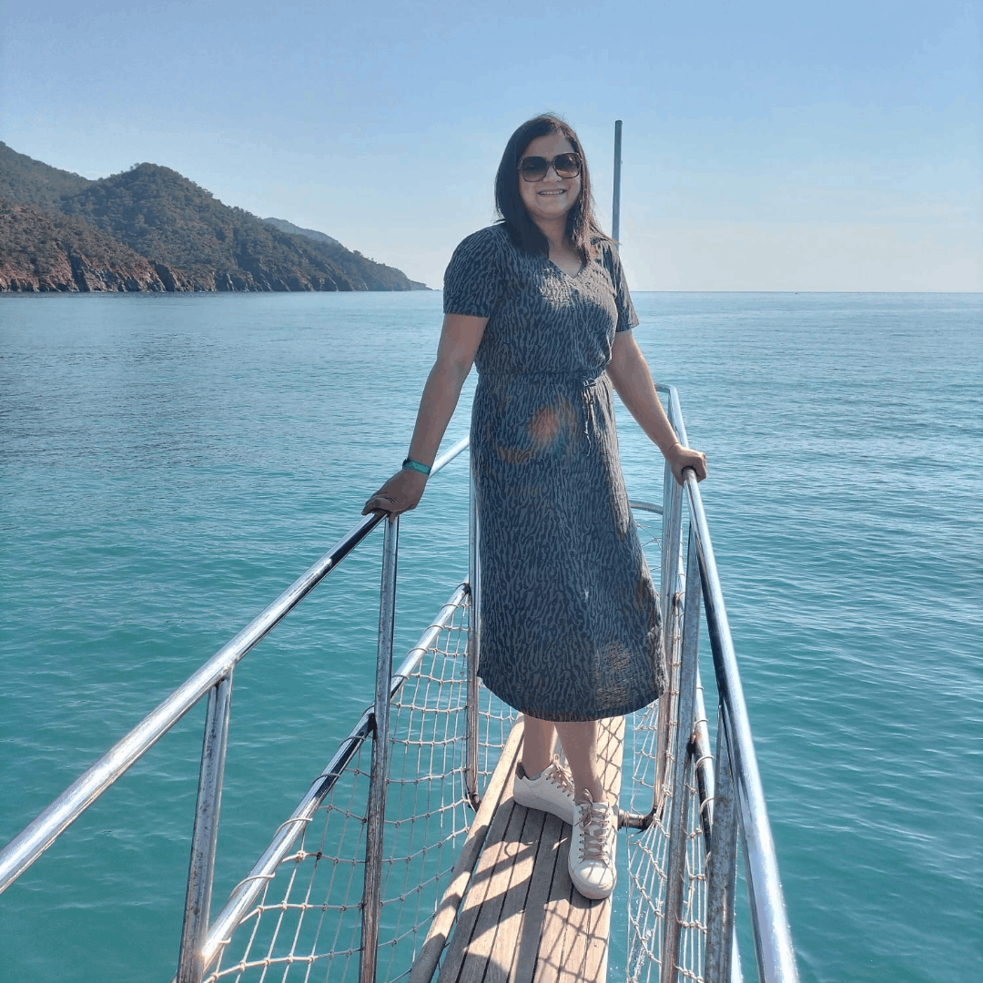 A woman in a patterned midi dress and white leather sneakers standing on the gangway of a boat on clear blue water.
