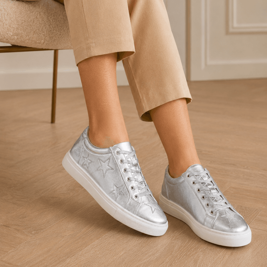 A person wearing Cocorose London Hoxton metallic silver low-top trainers with embroidered star details and white soles, paired with cropped beige trousers.