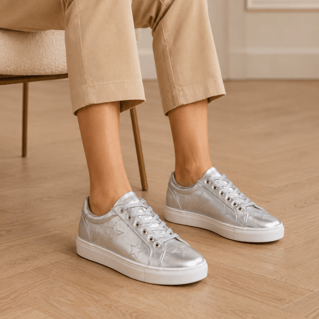 A person is seated in a modern interior showcasing their metallic silver leather trainers with stars and white soles by Cocorose London on a light wood floor.