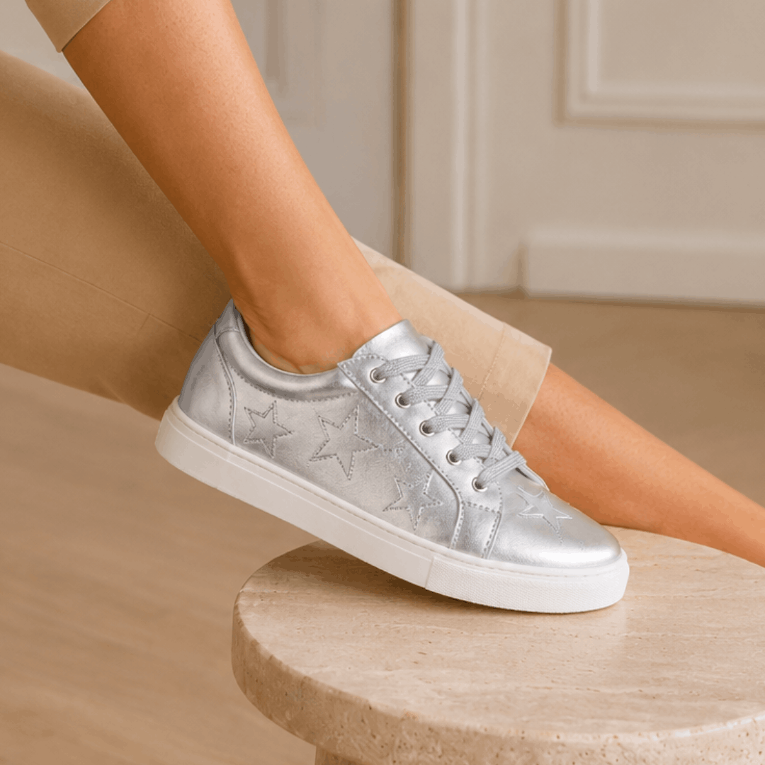 A side-profile view capturing a person's metallic silver low-top trainers with silver stars, silver laces and white soles resting on a stone stool in a contemporary interior setting.
