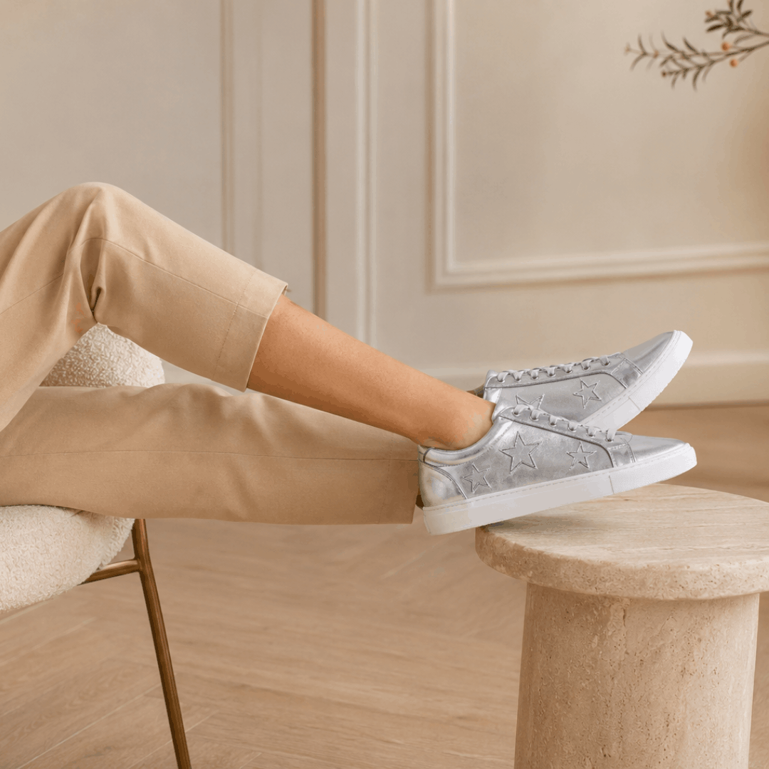 The image shows a relaxed, elegant perspective of a person resting their legs on a stone plinth while wearing silver leather Cocorose London star trainers with white soles.
