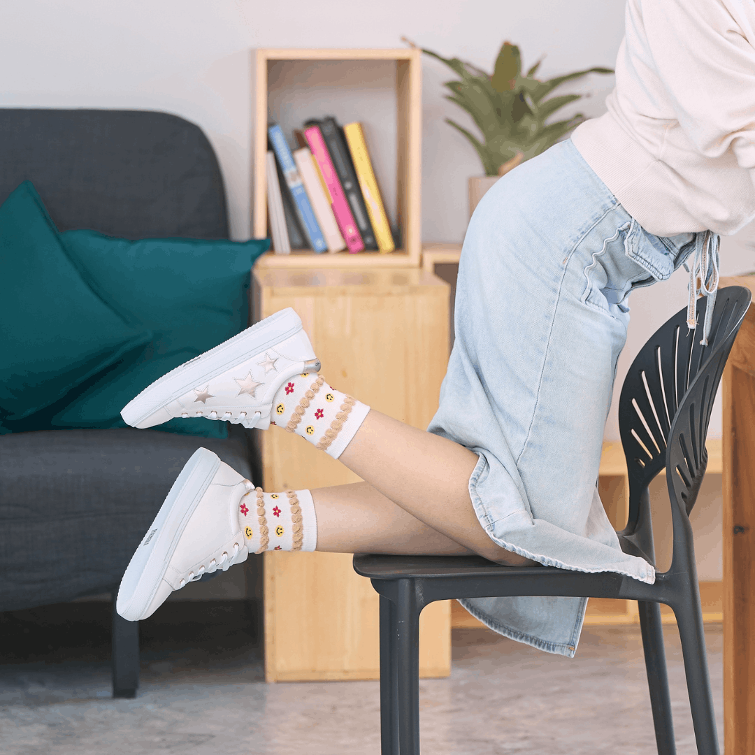 Side profile of a person kneeling on a chair wearing white leather trainers with gold star details on the outer side and metallic gold heel tabs, styled with pattern socks and a denim skirt.