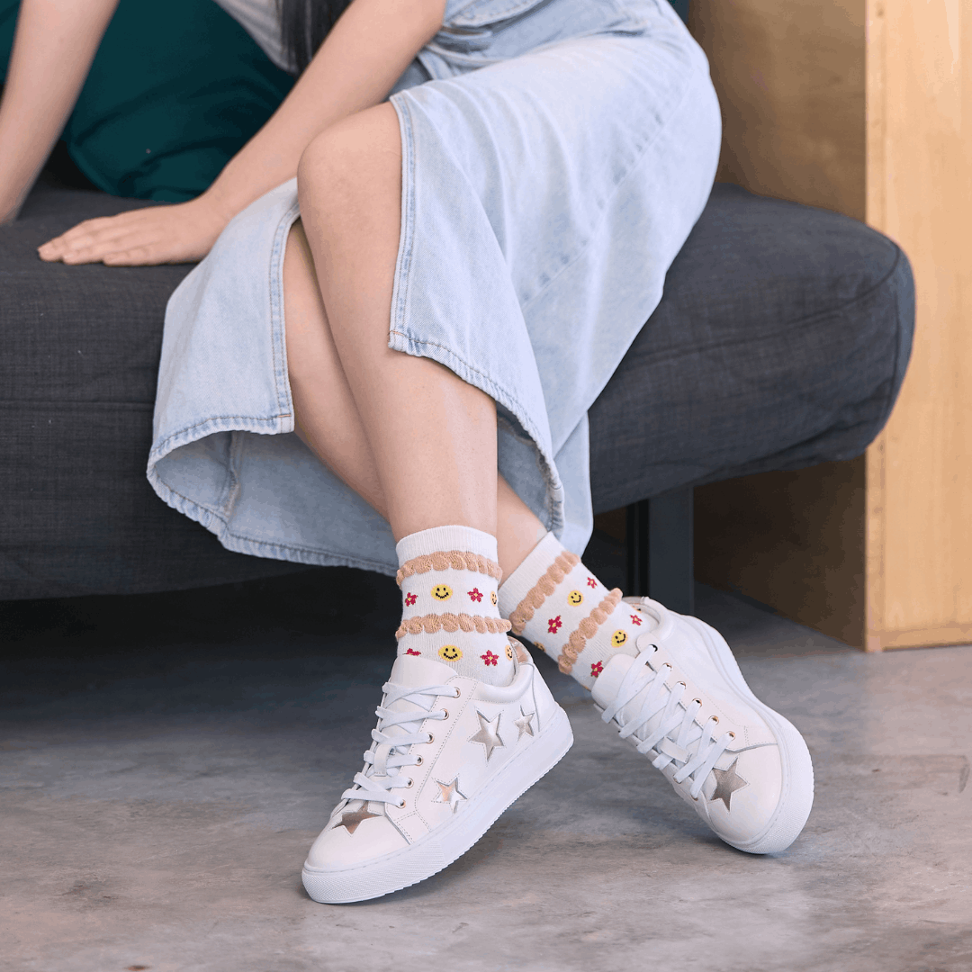 Front view of a person sitting on a dark grey sofa wearing white leather trainers with gold star appliqués on the side and toe, paired with patterned socks and a denim midi skirt.