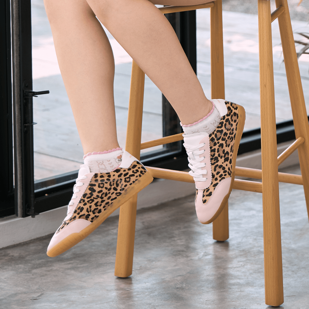 Person wearing leopard print shoes with pastel pink trim details and white laces on a wooden stool.