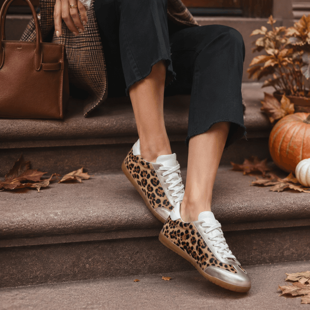 Close-up of a person sitting on stone steps wearing black trousers and Moorgate leopard print sneakers with gold trim next to a brown leather handbag and pumpkins.