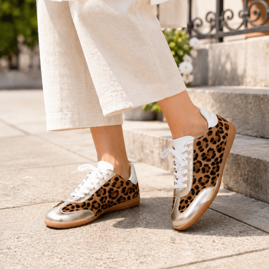 A person wearing cream wide-leg cropped trousers and Moorgate leopard print sneakers with gold details walking on a sunny stone pavement.