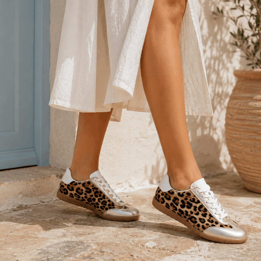 Side view of a person walking on a rustic stone patio wearing a white dress and Moorgate leopard print trainers with metallic gold accents.