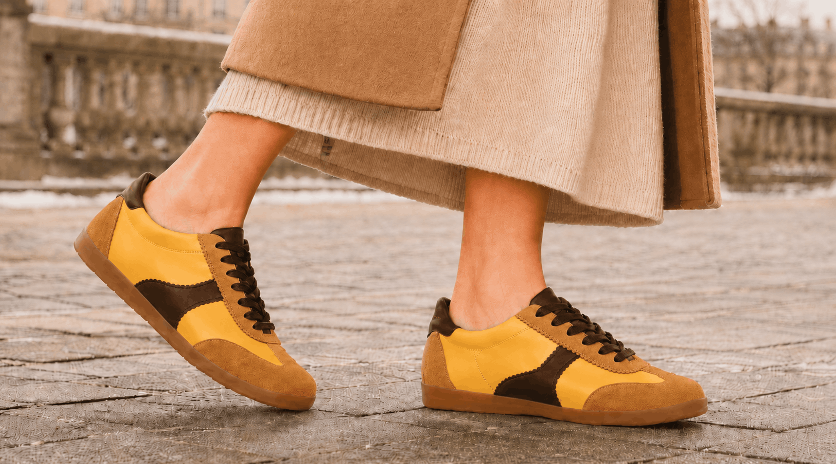 Close-up of a person walking on a cobblestone path wearing yellow and chocolate brown leather trainers paired with a beige knit skirt.