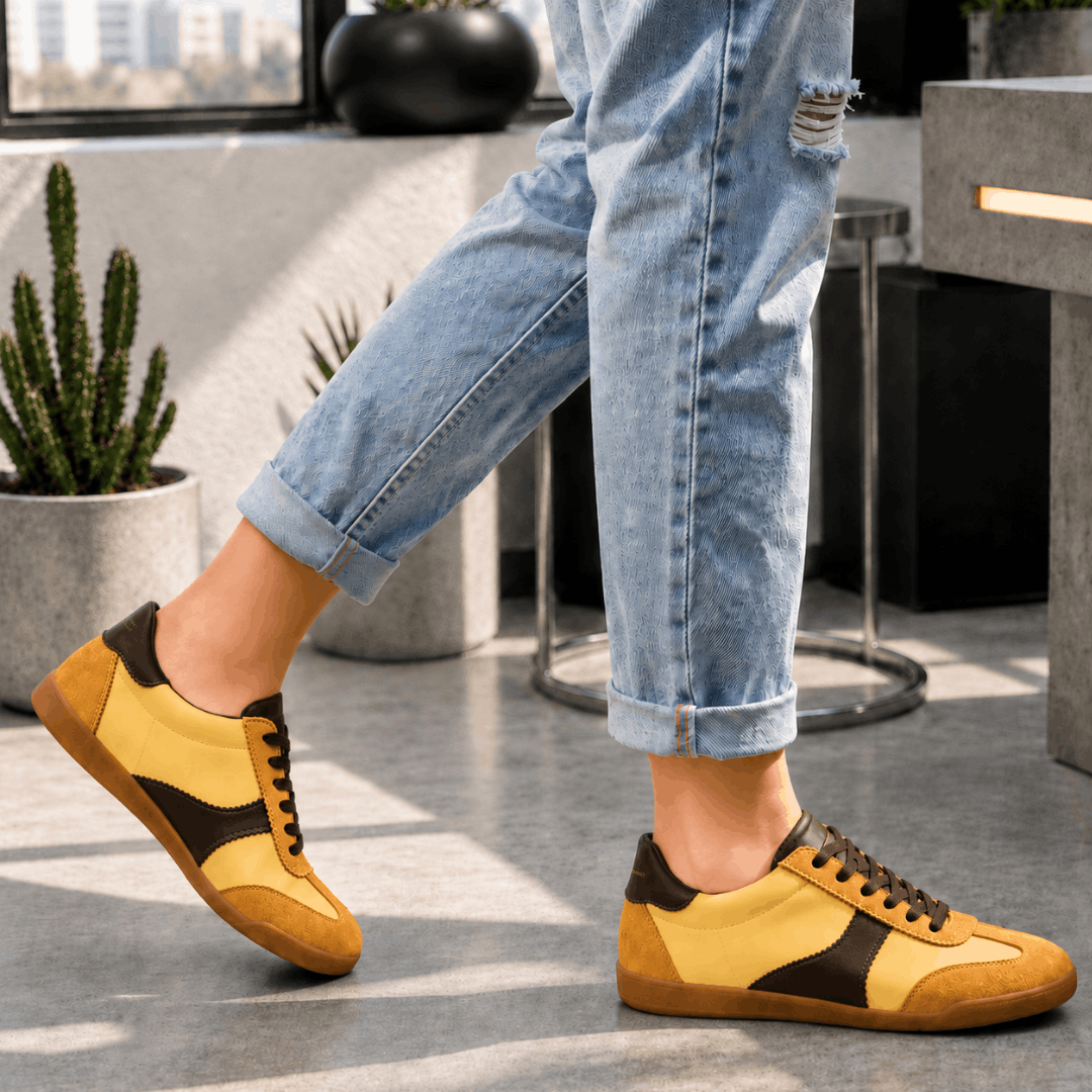 A side-profile view of a person walking in light-wash, cuffed jeans and wearing yellow and chocolate brown low-top leather trainers
