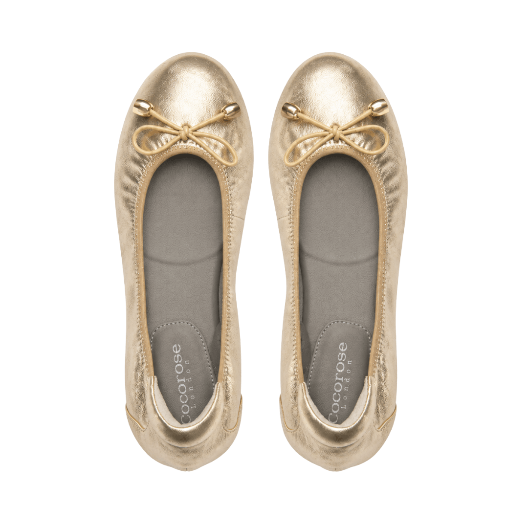 A direct top-down, bird's-eye view of a pair of metallic gold ballet flats with beige bow details and gray insoles printed with "Cocorose London," isolated on a white background.