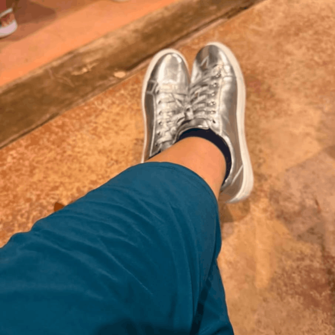 Point-of-view shot looking down at crossed legs wearing blue trousers and metallic silver low-top lace-up trainers with white soles.