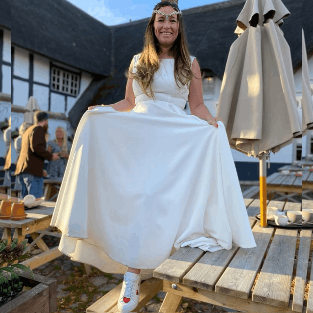 Bride smiling outdoors in a sleeveless white wedding gown, lifting the hem to show white leather trainers with red heart detailing.