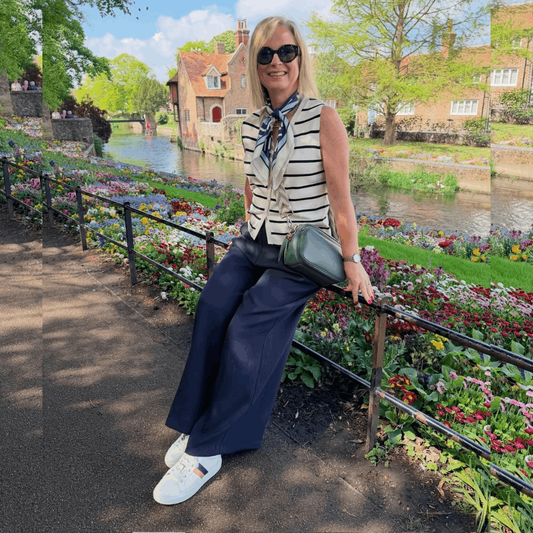 Woman sitting on a park railing along a scenic canal, wearing navy wide-leg trousers, a striped top, and white leather sneakers.