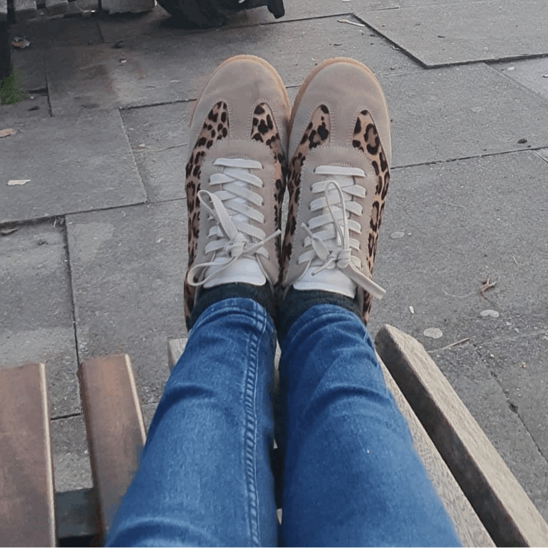 Top-down view of a person seated outdoors wearing blue jeans and suede low-top sneakers with leopard print side panels.