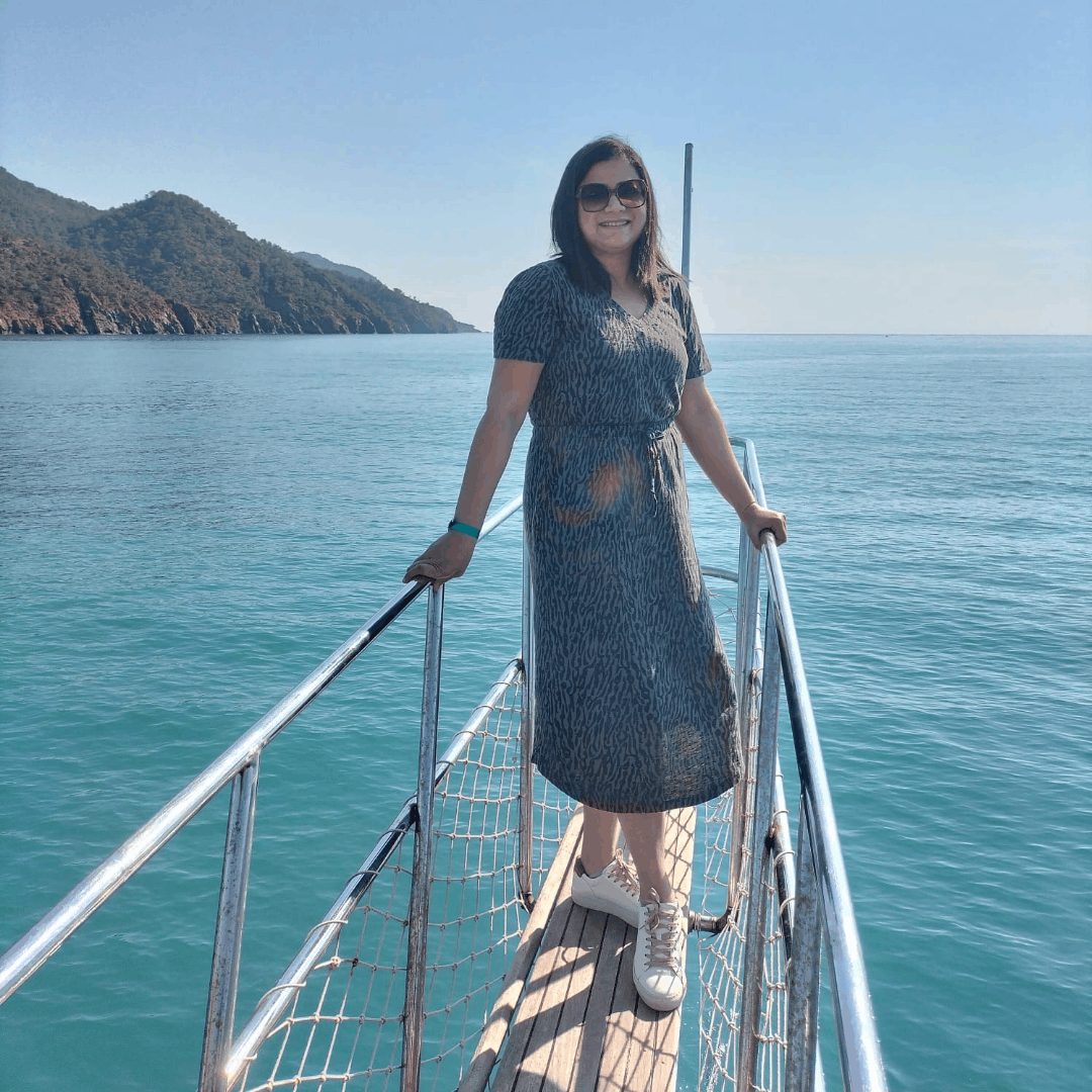 Woman in a grey patterned midi dress and white leather trainers standing on the bow wooden deck of a boat in turquoise water.