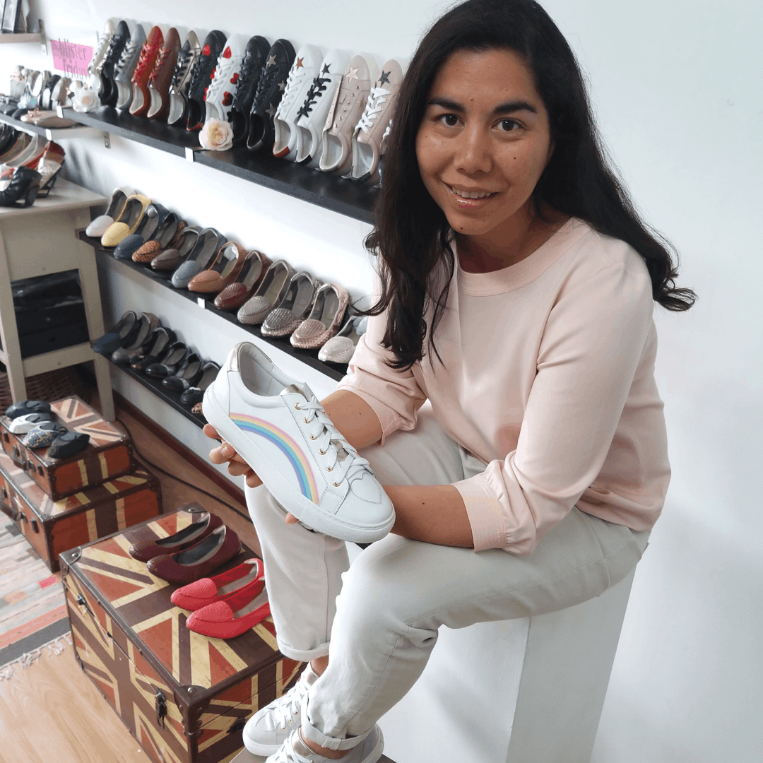 Janan in Cocorose Studio holding a white leather trainer with a rainbow side stripe, surrounded by displays of shoes and Union Jack trunk tables.