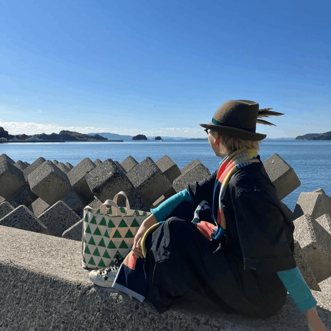 A person sits outdoors on a concrete seawall overlooking the ocean while wearing a casual outfit and Cocorose London trainers.