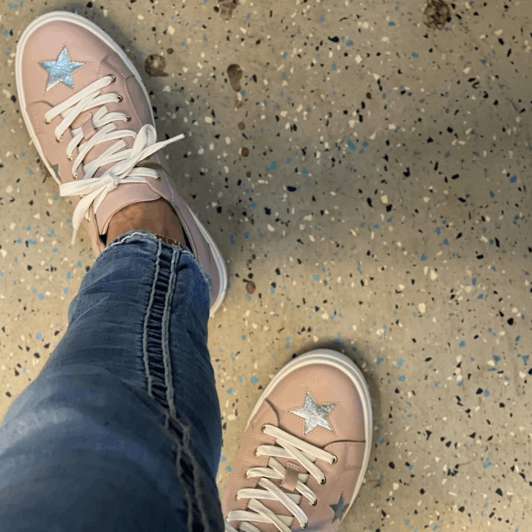 A top-down view captures a person's feet in pastel pink Cocorose London leather trainers adorned with vibrant metallic blue embroidered stars.